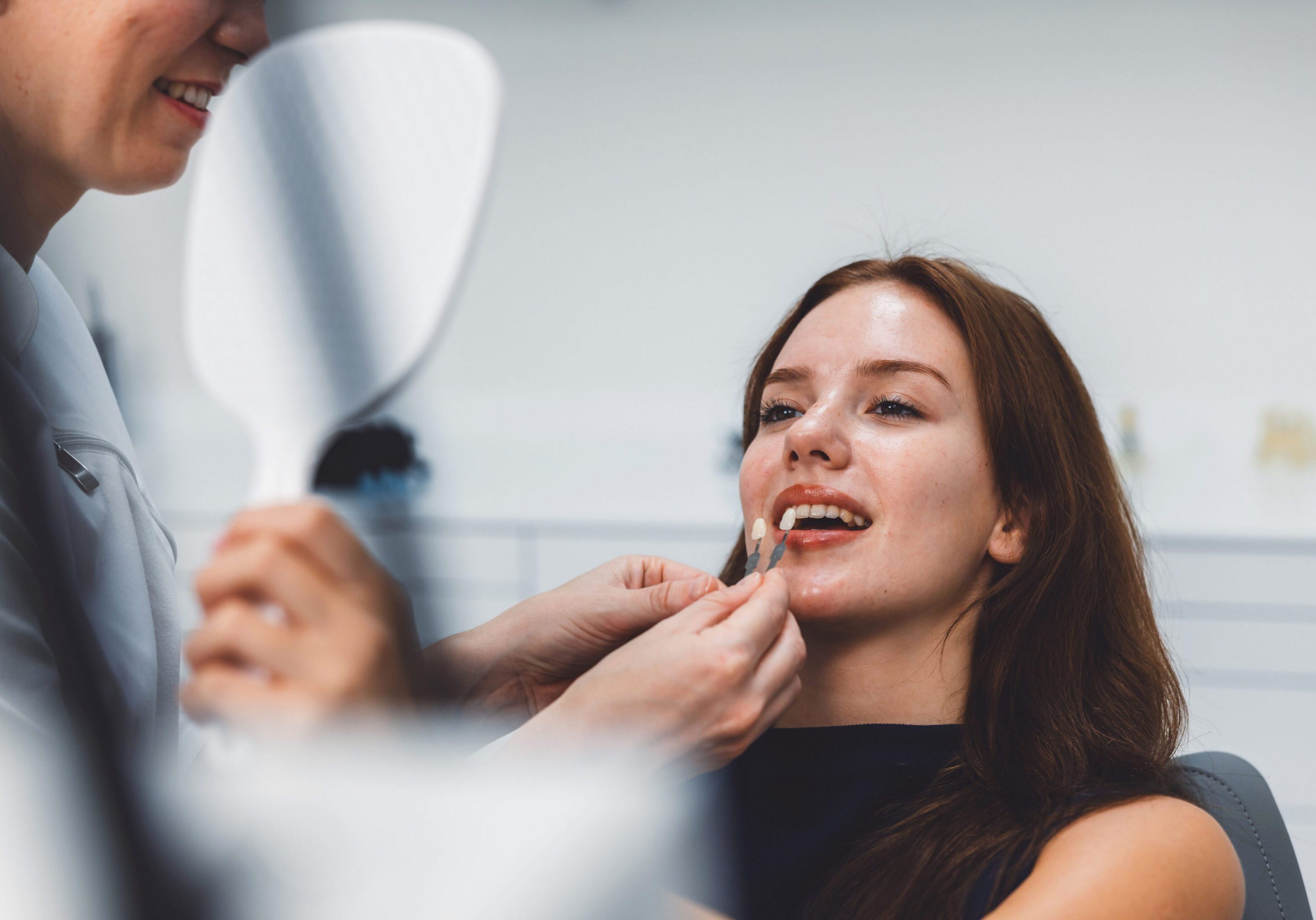 A dental professional is fitting a dental veneer on a smiling patient in a modern dental clinic. The patient looks pleased as she examines her smile in a handheld mirror.